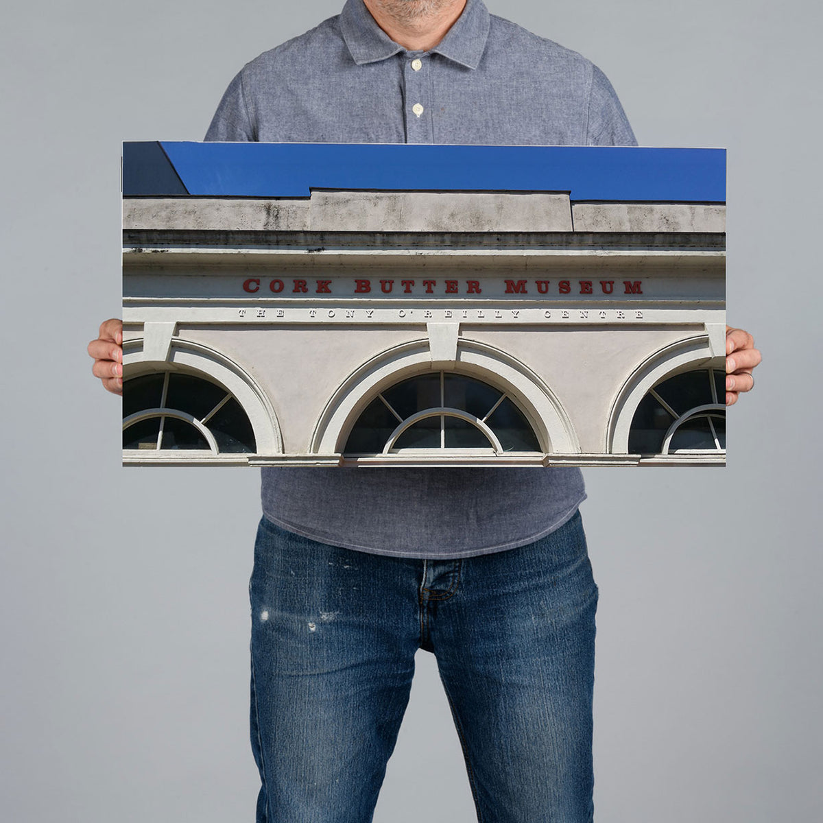 A man holds a 60x30cm panoramic canvas print art featuring the facade of Cork Butter Museum. The print has a blue upper section and a light gray lower section with red lettering.
