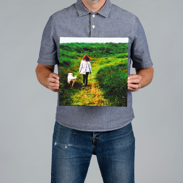 A man from The Canvas Works holds up a square 30x30cm canvas print showing a woman walking a dog on a grassy path. 