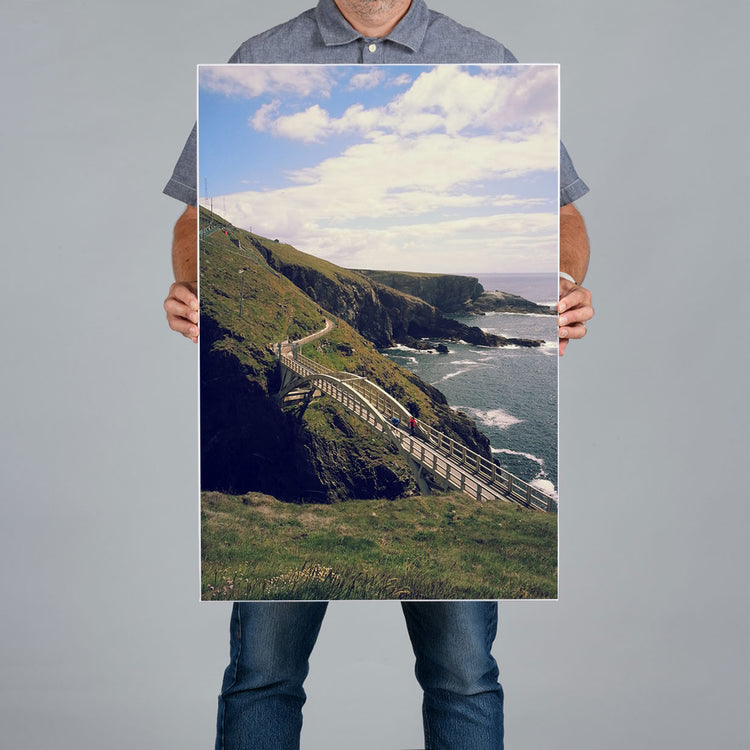 A person from The Canvas Works holding a large vertical50x75cm canvas print featuring a coastal landscape from The Mizen Head in Cork with grassy cliffs, a winding pathway, and the ocean under a cloudy sky, against a plain light gray background.