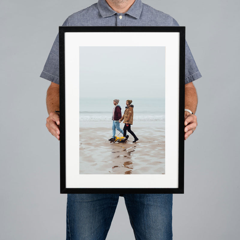 A man holds up a 30x45cm black framed art print against a solid gray background. The print features a photograph of a couple walking on a beach, framed with a white mat and a black frame.