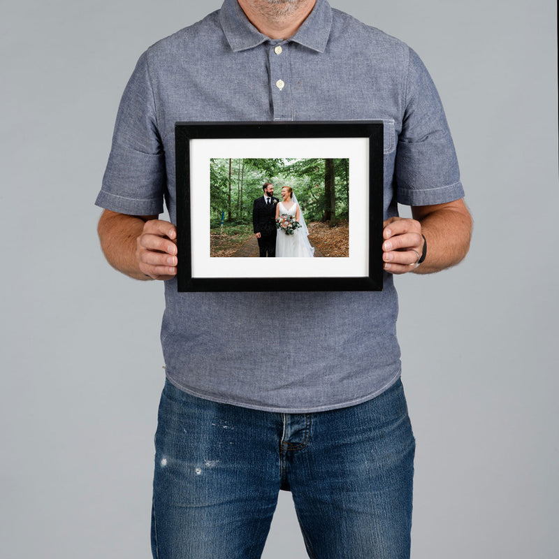 A man holds a 20x15cm framed photo print against a solid gray background. The frame is black with a white mat, showcasing a custom wedding photograph of a couple in a forest.